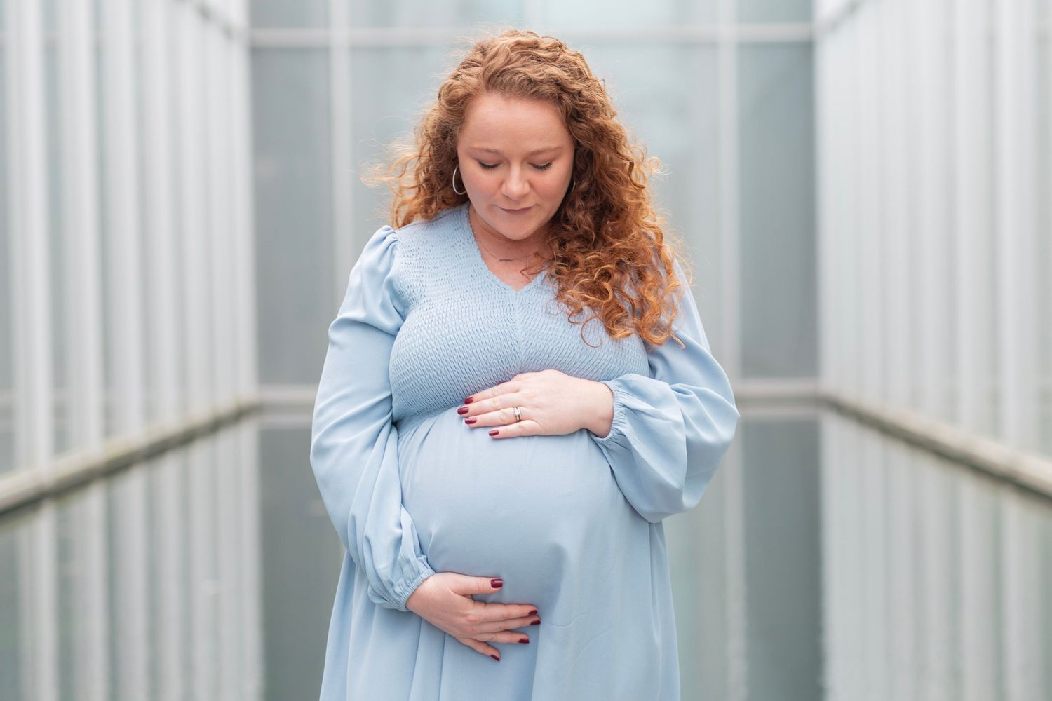 Expectant mother in soft blue dress cradles her baby bump while standing in a outdoor corridor at the NC Museum of Art in Raleigh.
