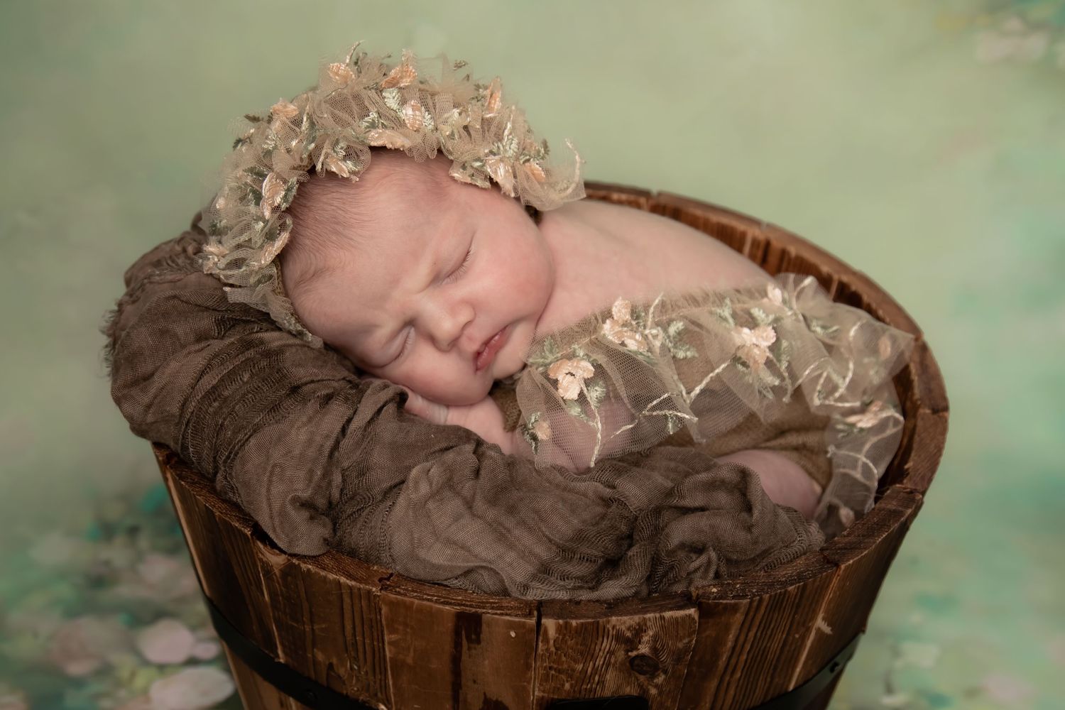 Newborn sleeping in rustic wooden bucket with floral crown against mint green backdrop.