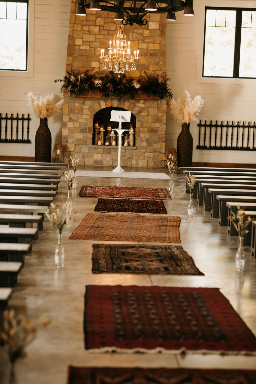 Rustic wedding venue interior with Persian rugs lining aisle, stone fireplace, and chandelier altar.