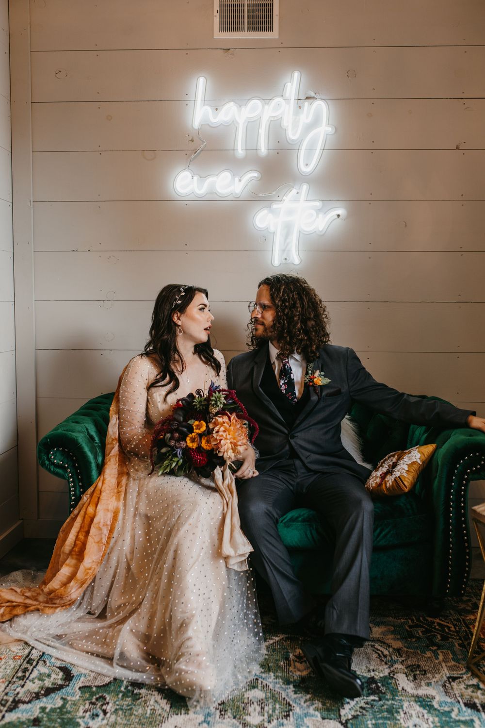 Bride and groom sit on green velvet sofa beneath glowing happily ever after neon sign.