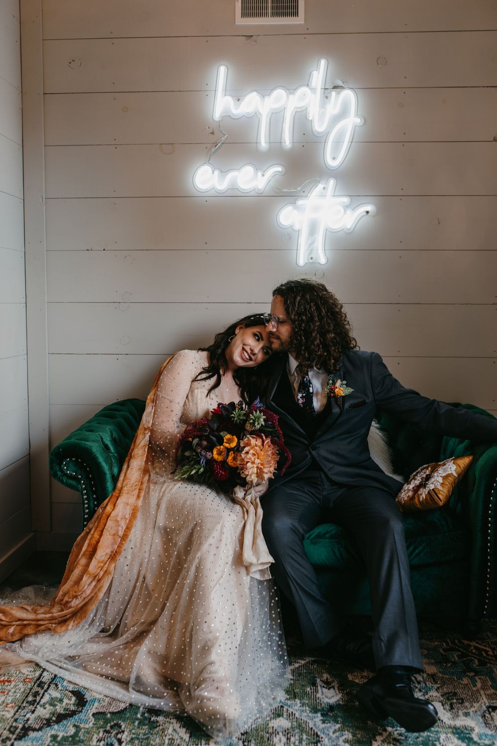 Newlyweds share tender moment on emerald velvet sofa under happily ever after neon wedding sign.