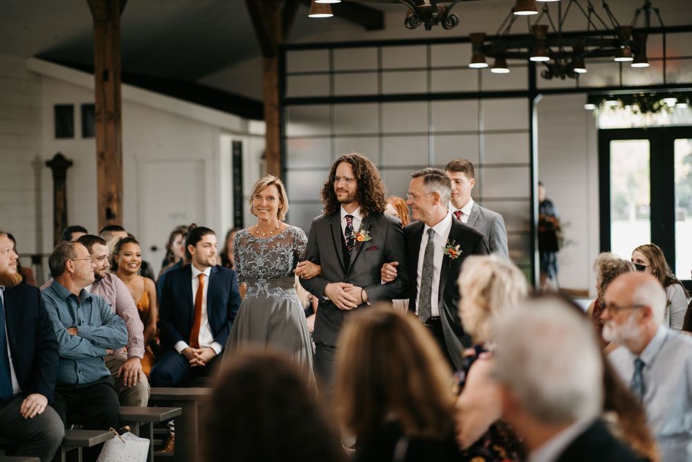 Groom walks down wedding ceremony aisle escorted by parents past seated guests.