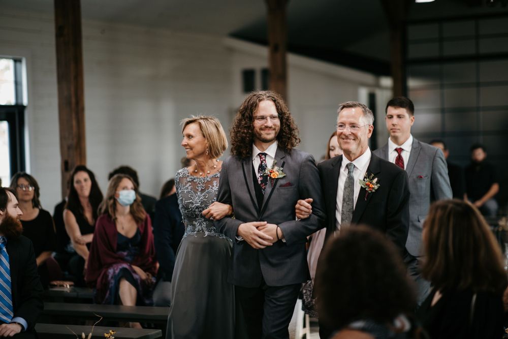 Groom in dark suit walks down aisle flanked by proud parents during wedding ceremony.