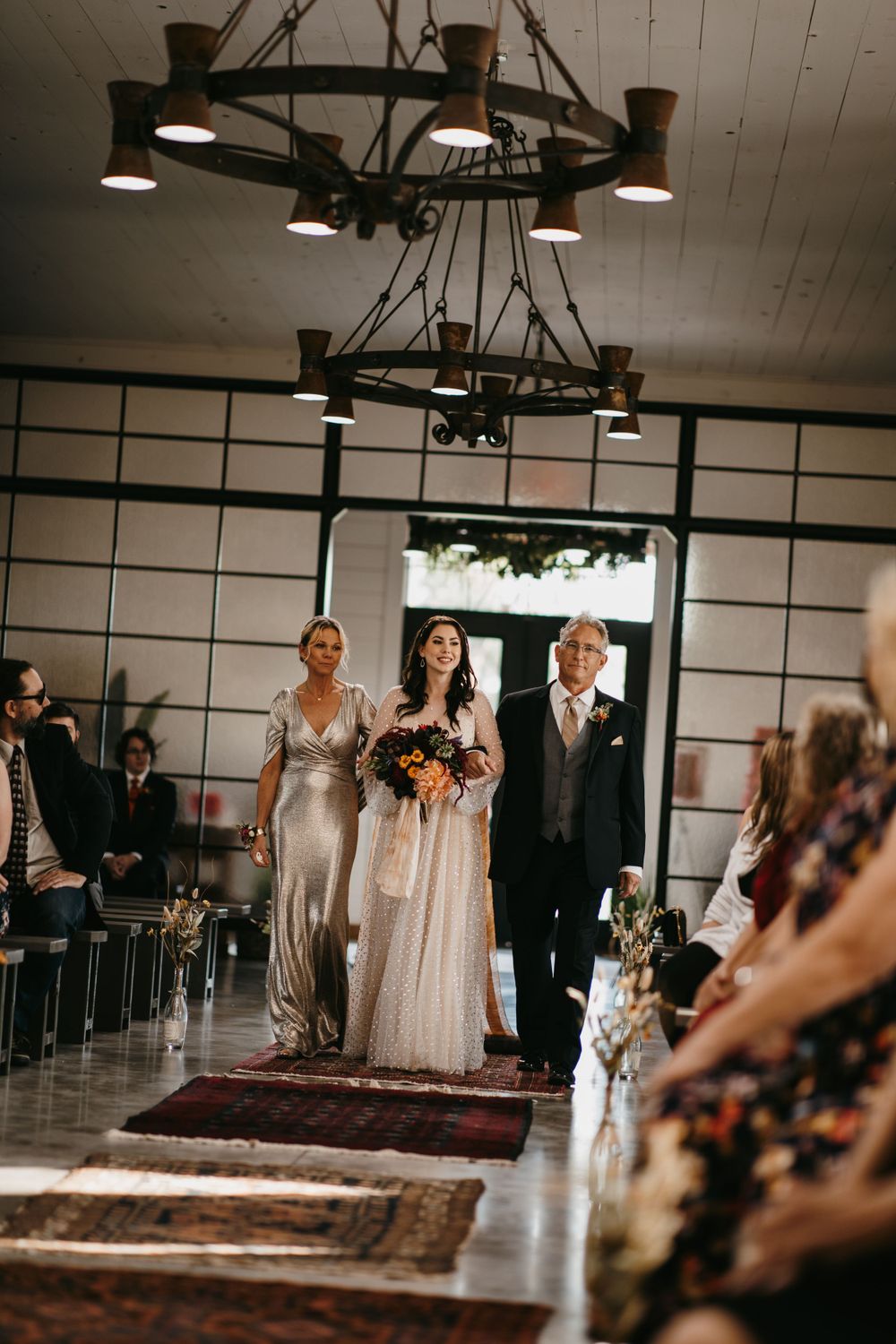 Bride walks down rustic venue aisle with parents, carrying vibrant autumn bouquet past guests.