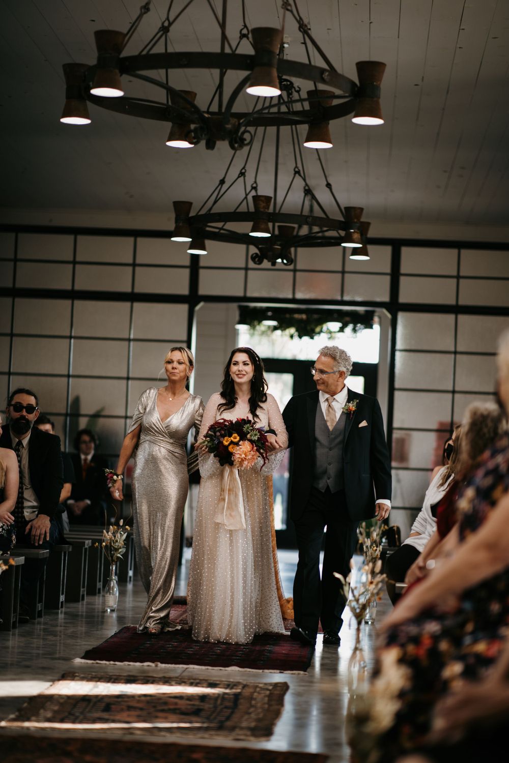 Bride walks down the aisle flanked by parents in elegant wedding venue with rustic chandelier.