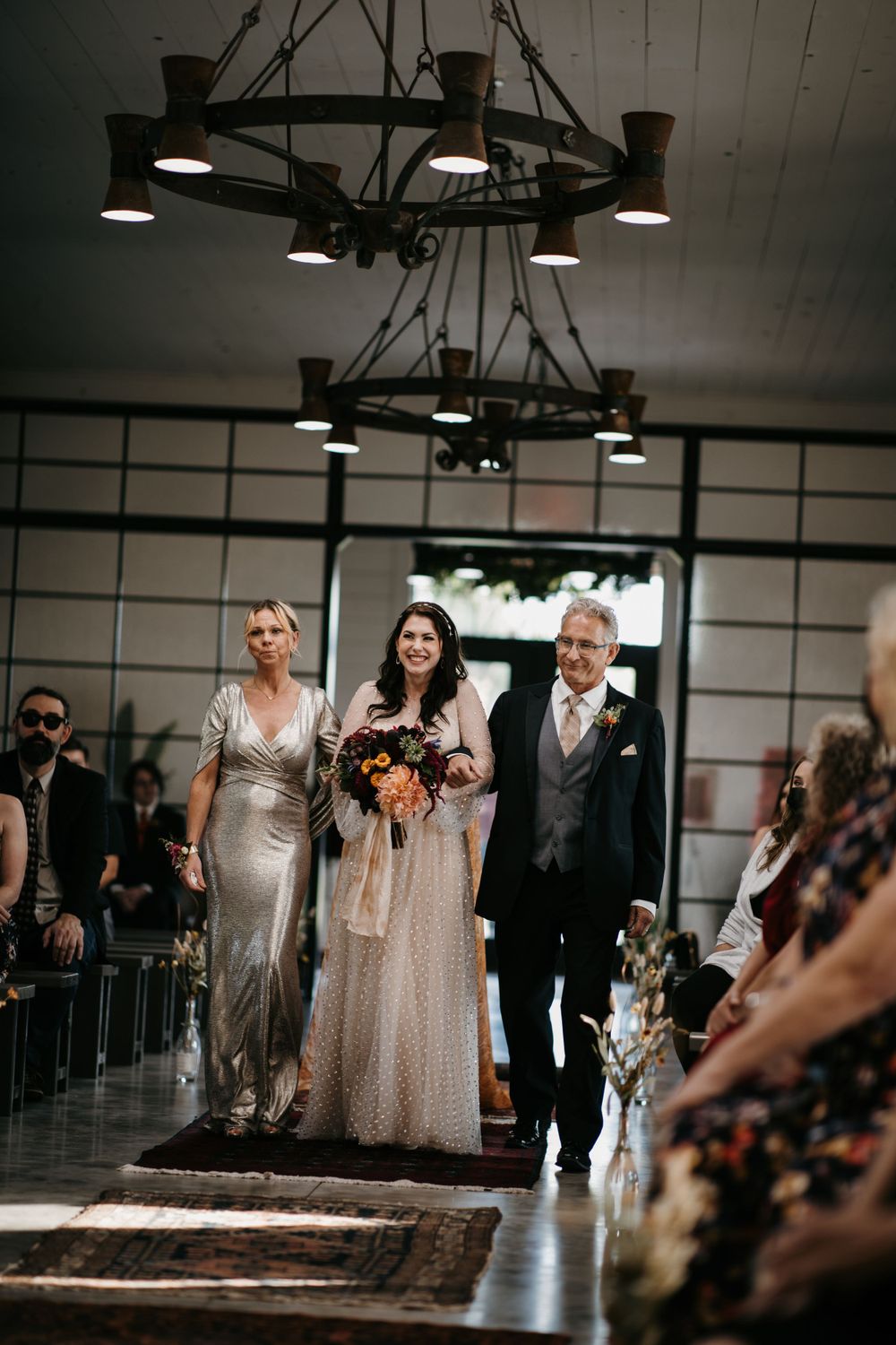 Bride in champagne gown walks down aisle with parents under rustic chandelier at wedding ceremony.