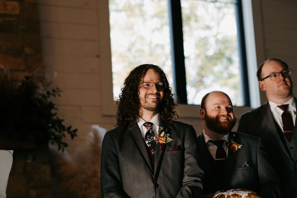 Emotional groom smiles watching bride walk down aisle at indoor wedding ceremony.