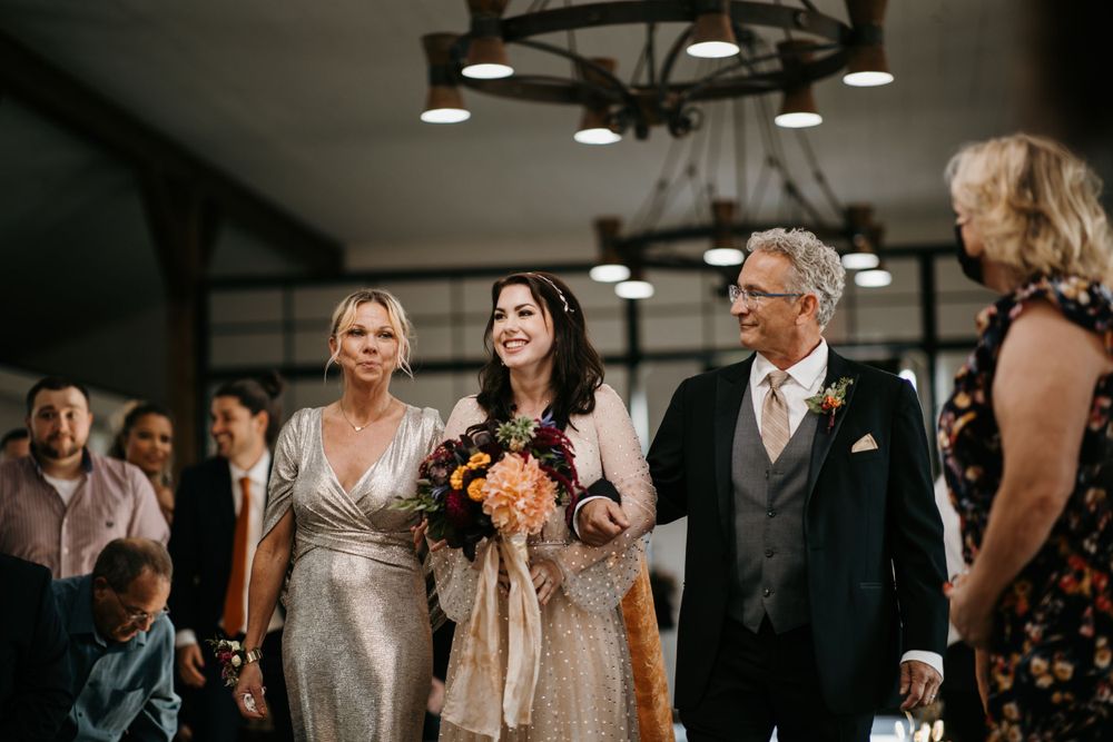 Bride walks down wedding aisle with parents, holding colorful bridal bouquet in barn venue.