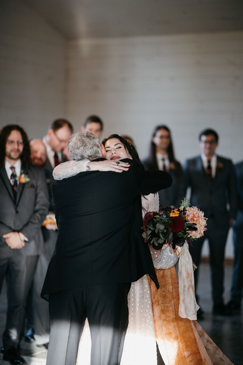Bride embraces father in emotional moment at wedding ceremony altar with wedding party.