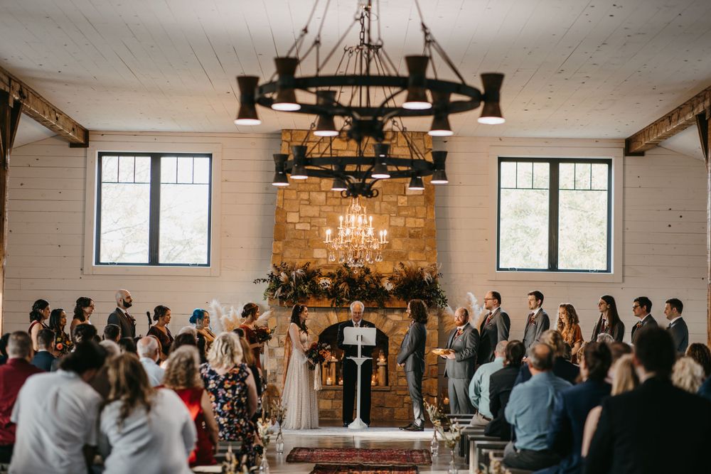 Wide shot of rustic barn wedding ceremony with stone fireplace and chandelier backdrop.