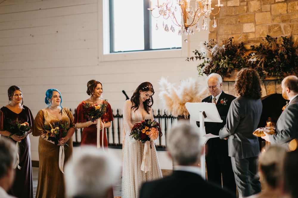 Bride stands at altar with bridesmaids in jewel-toned dresses during rustic barn ceremony.