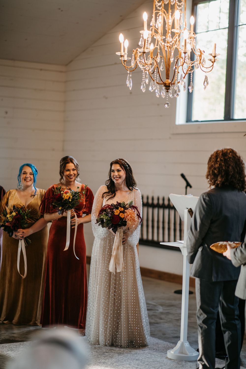 Bride and bridesmaids holding colorful bouquets stand at altar in white barn wedding venue.