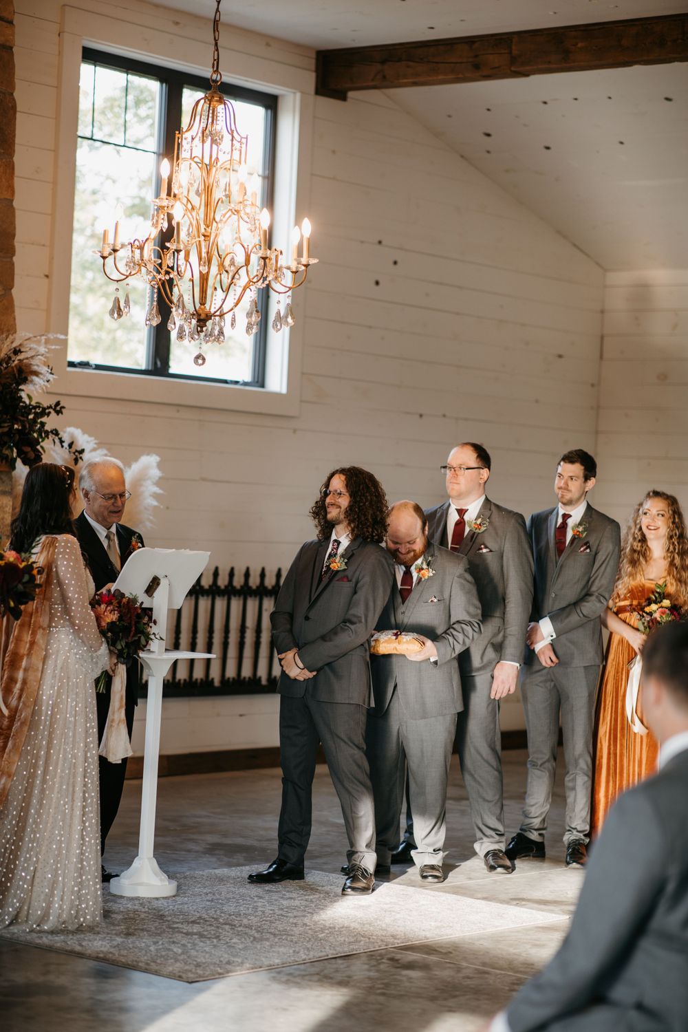 Groom and groomsmen in gray suits stand at altar during rustic barn wedding ceremony.
