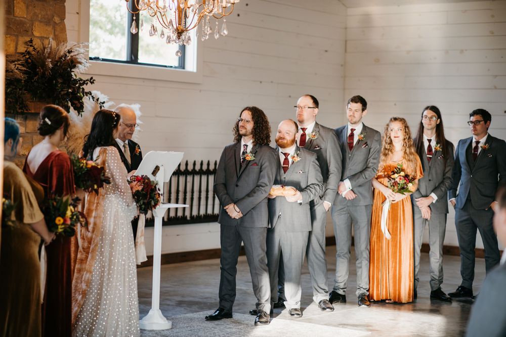 Wedding party stands at altar in white barn venue with crystal chandelier overhead.