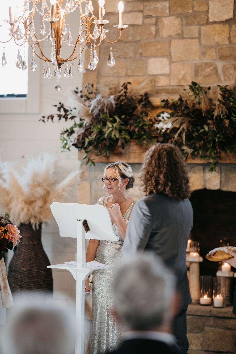 Officiant reads vows at white podium during indoor wedding ceremony with stone fireplace and crystal chandelier.
