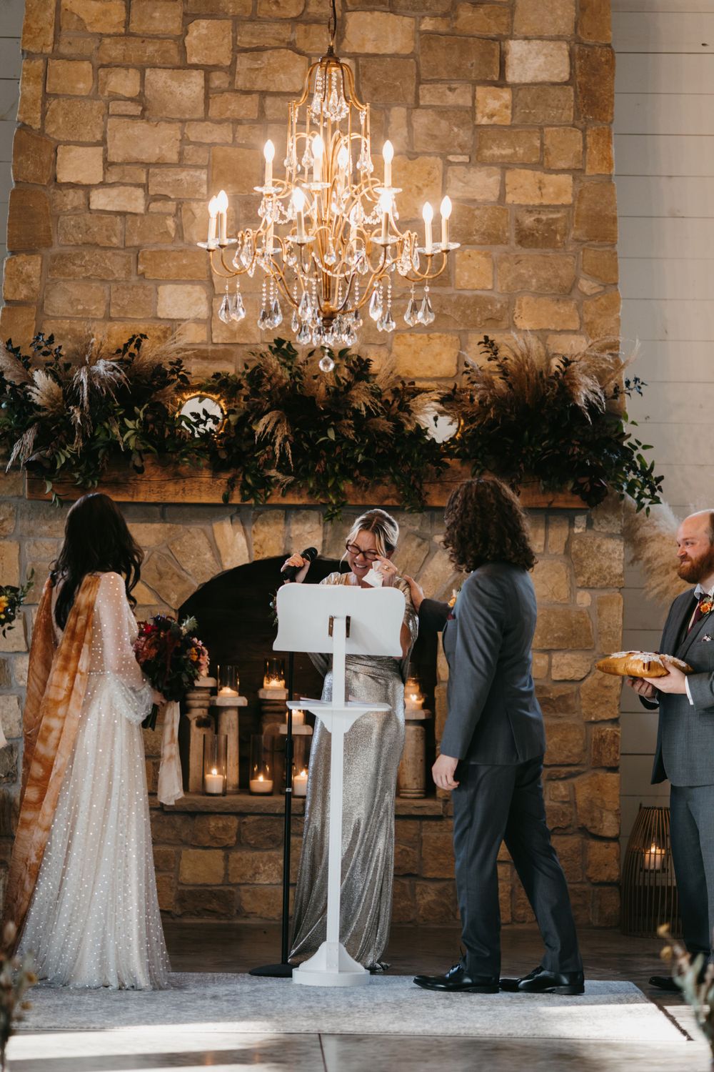Wedding ceremony at stone fireplace altar with crystal chandelier, candles, and lush greenery garland decor.