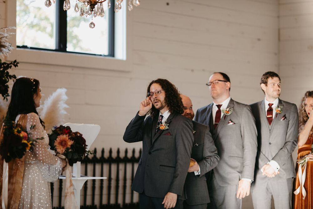Emotional groom wipes tears during wedding ceremony beside groomsmen in gray suits with floral boutonnieres.