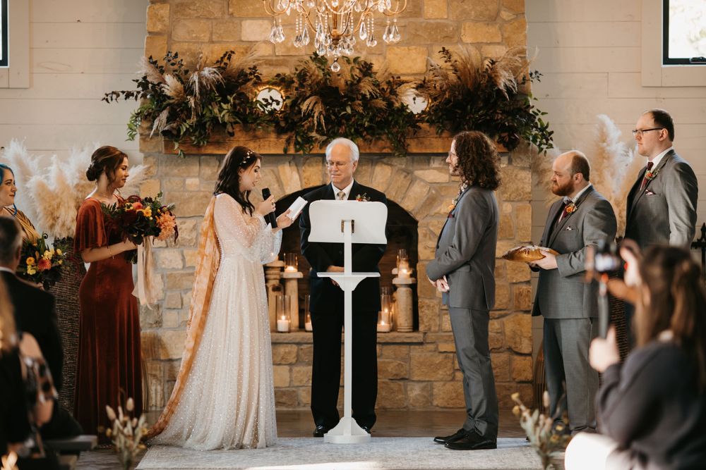 Bride and groom exchange vows at stone fireplace altar with elderly officiant and crystal chandelier above.