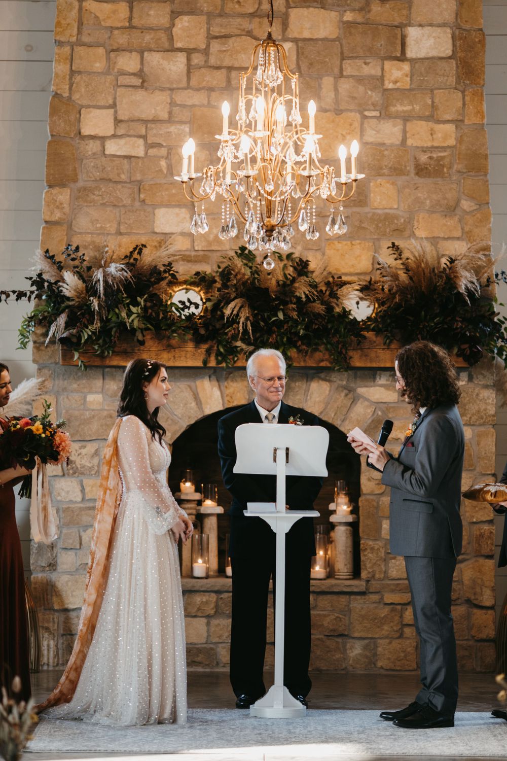 Bride and groom share vows at stone fireplace altar with crystal chandelier and greenery garland backdrop.