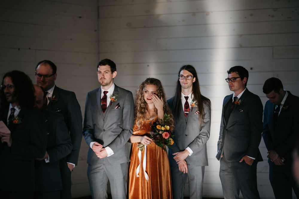 Bridesmaid in orange dress wipes tears while standing with groomsmen in gray suits during wedding ceremony.