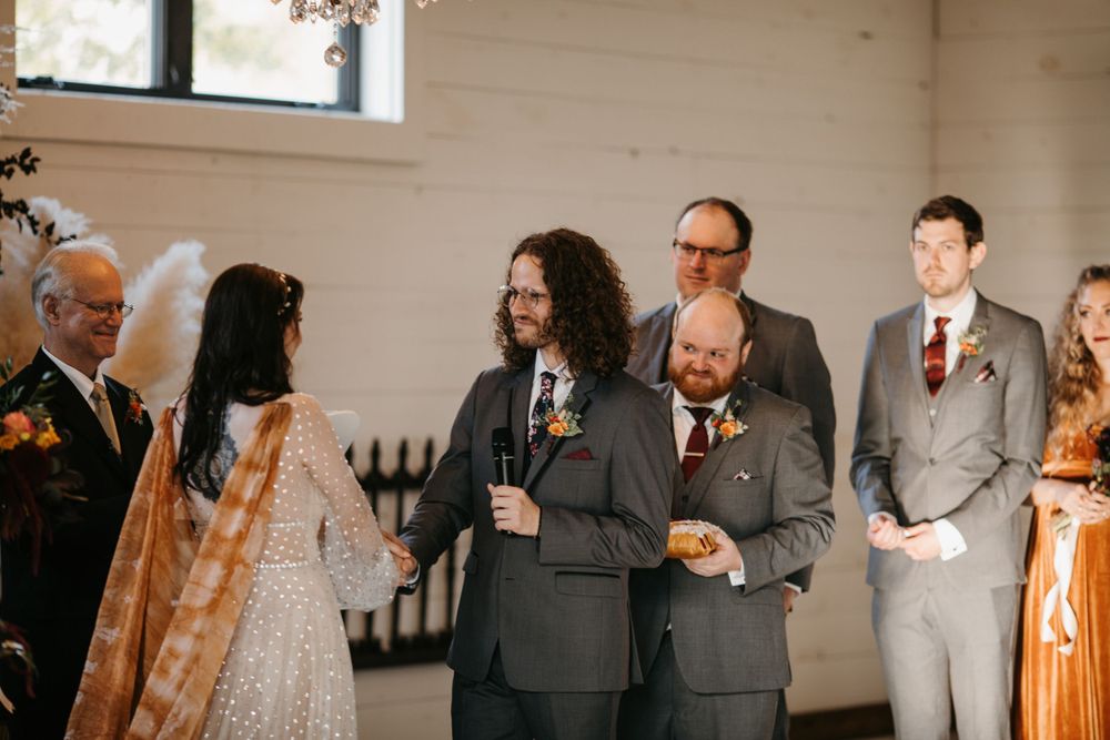 Groom reads vows while wedding party stands beside stone fireplace altar during indoor ceremony.