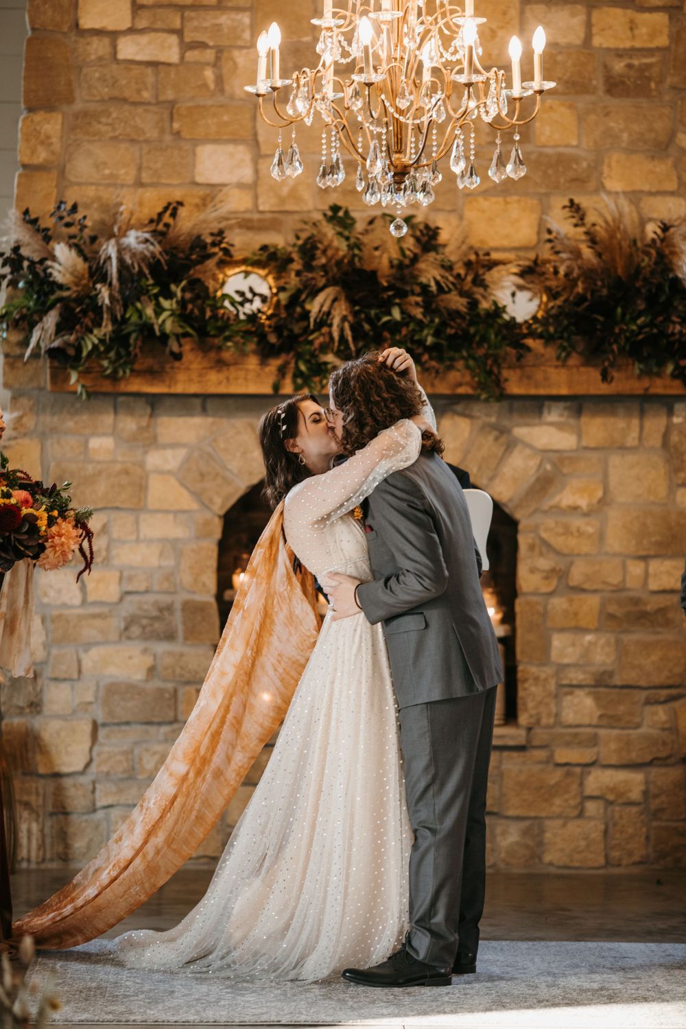 Bride and groom share first kiss at stone fireplace altar beneath crystal chandelier at wedding ceremony.