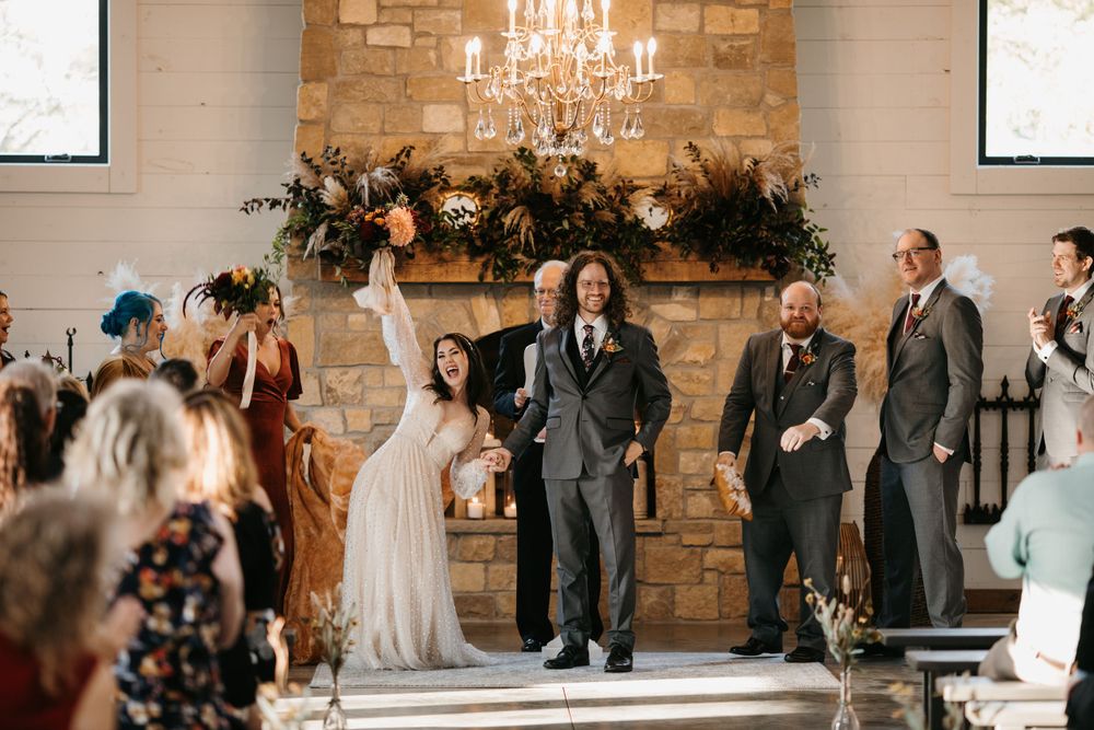 Newlywed bride raises bouquet in celebration while exiting stone fireplace altar after indoor wedding ceremony.