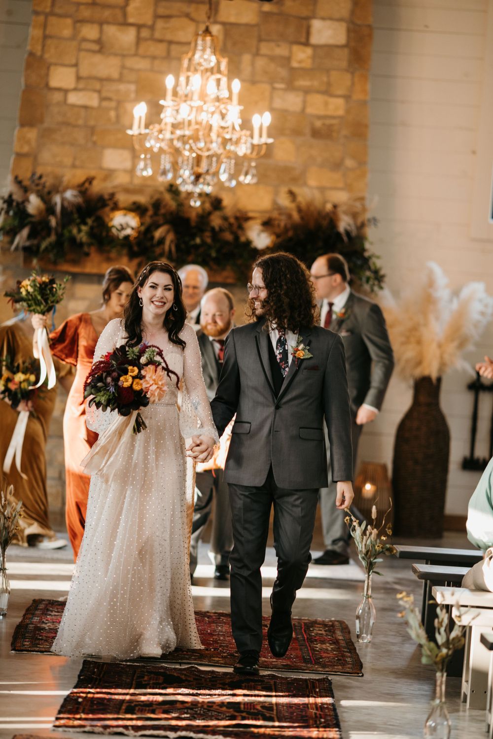 Smiling newlyweds walk down aisle hand in hand after wedding ceremony in rustic stone venue.
