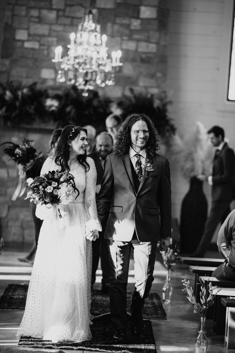 Bride and groom walk down the aisle smiling after wedding ceremony in rustic stone chapel with chandelier.