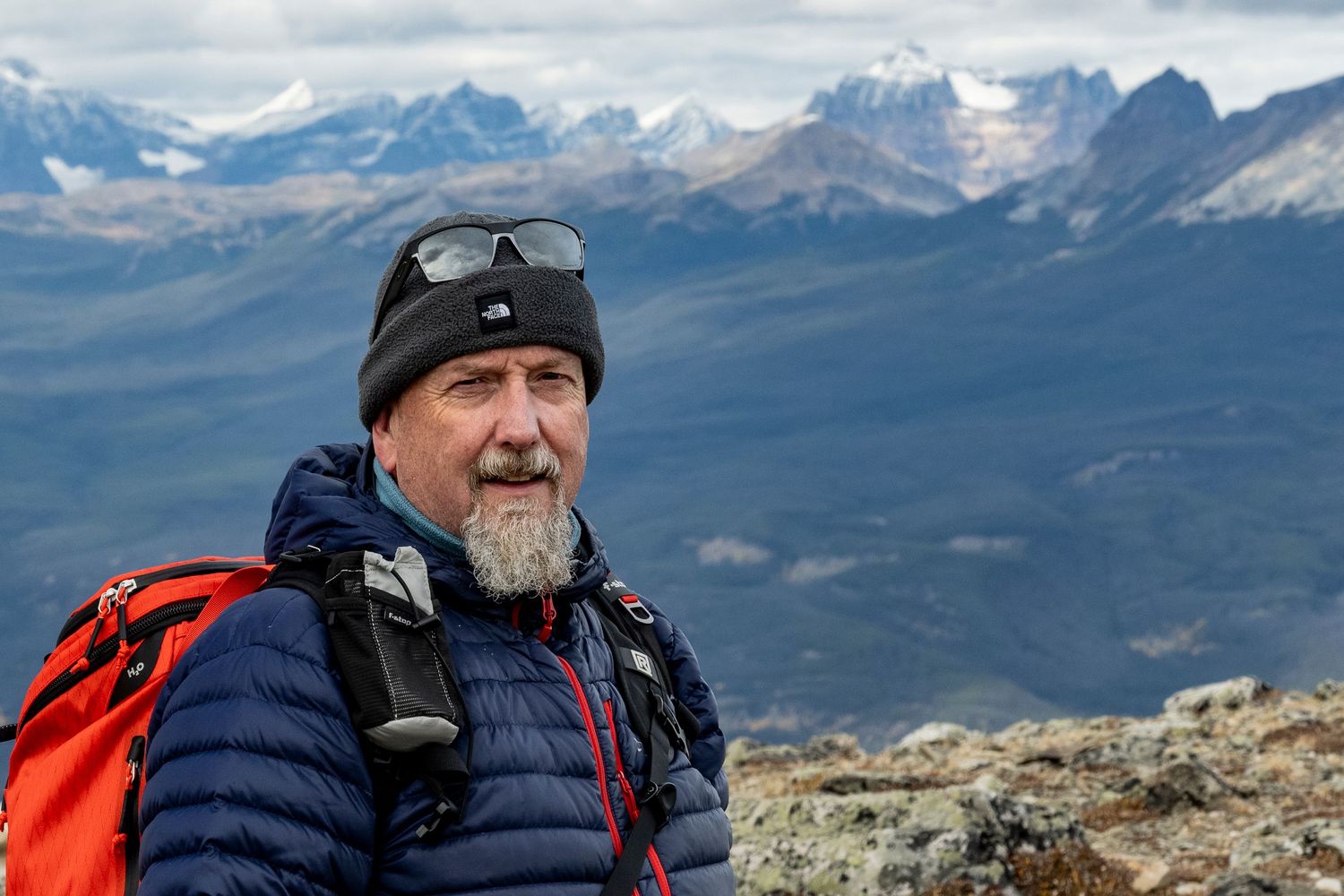 An adventurer wearing a winter hat and puffy jacket poses on a mountain summit with snowy peaks in the background.