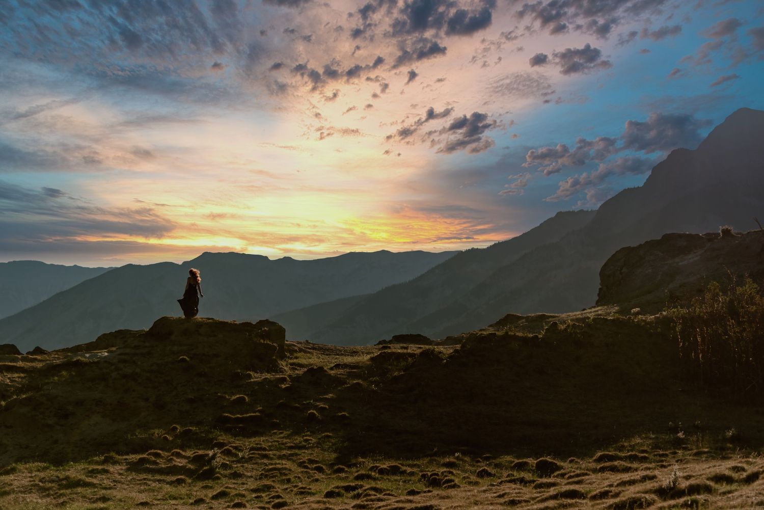 Silhouette of a woman in a long dress standing on rocky outcrop at sunset with dramatic cloudy sky over mountain landscape.