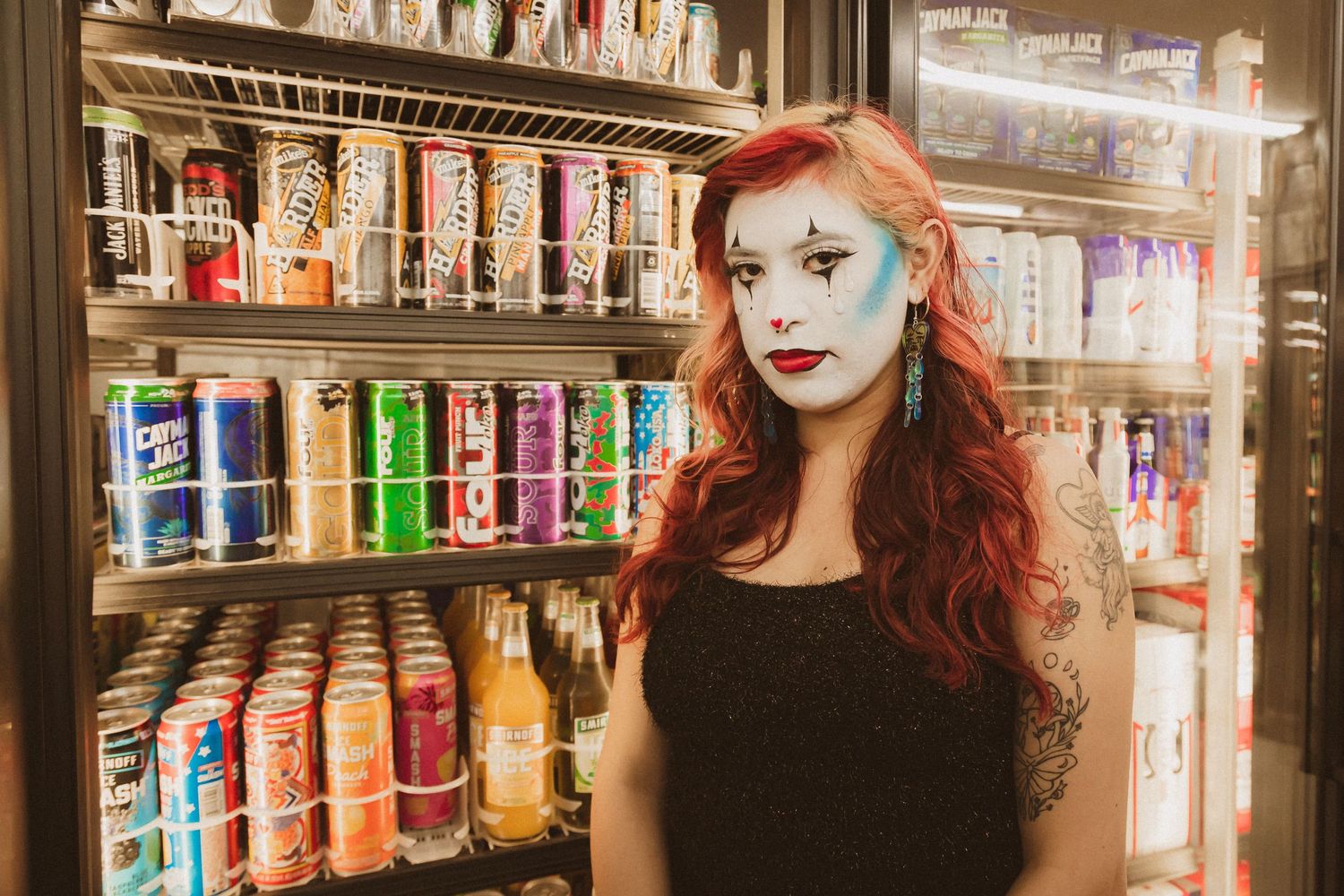 Person with red hair and face paint posing in front of colorful drink coolers.