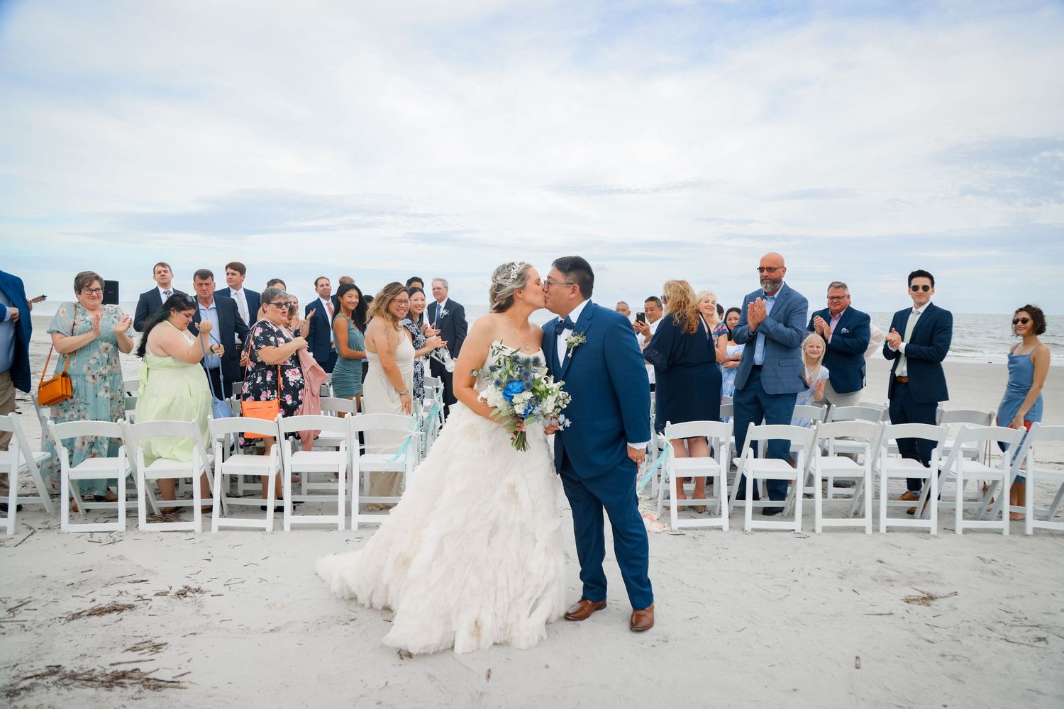 A beach wedding ceremony with guests seated in white chairs watching the couple exchange vows.