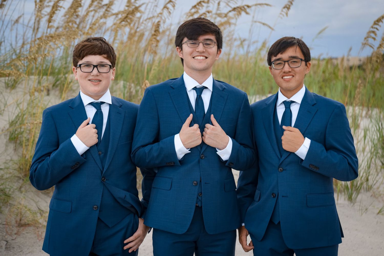 Three groomsmen in matching navy blue suits pose on a beach with sea oats.