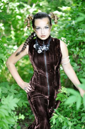 Model in brown velvet dress poses among green foliage with steampunk-inspired styling.