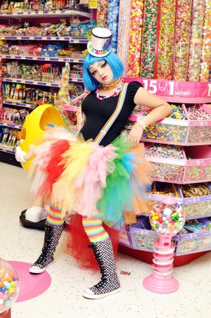 Colorful candy shop scene with person in rainbow tutu and blue wig surrounded by sweets displays.