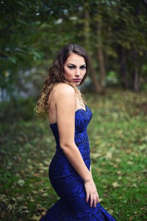 A woman in a royal blue velvet gown poses dramatically in a garden setting.