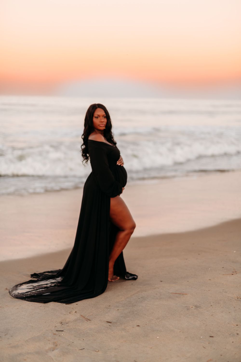 Expectant mother poses in black gown on beach with waves crashing behind.