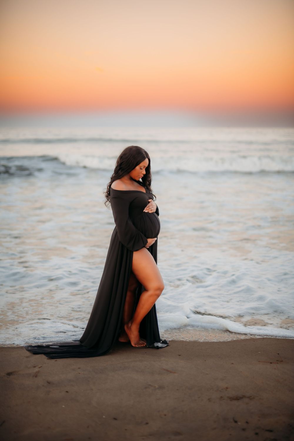 Maternity photoshoot on beach at sunset featuring elegant black flowing dress and golden sky.