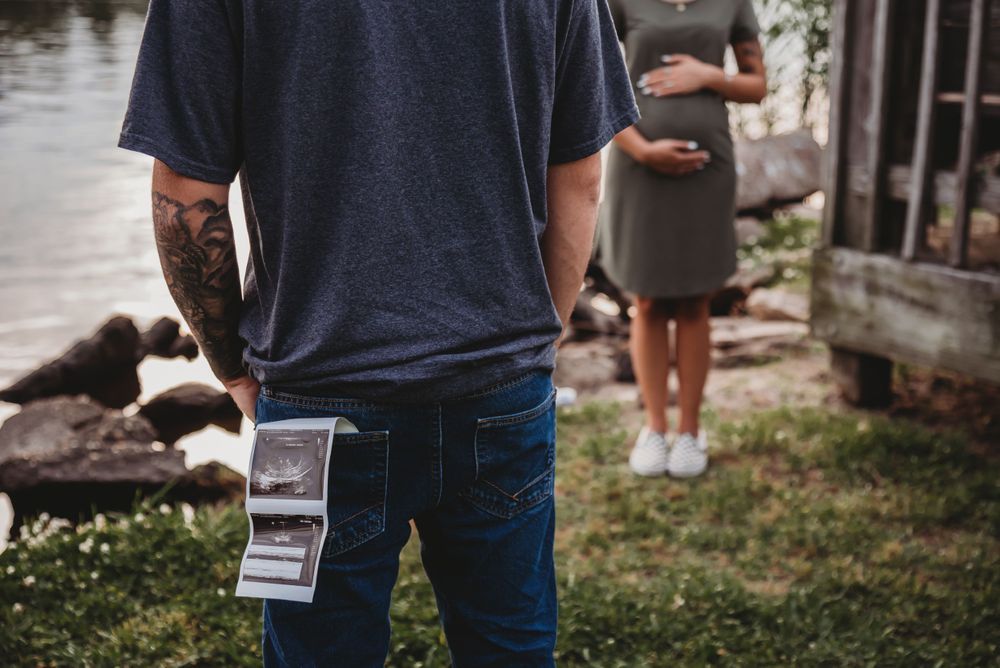 Tattooed person in casual attire holding ultrasound photos outdoors with someone standing in background.