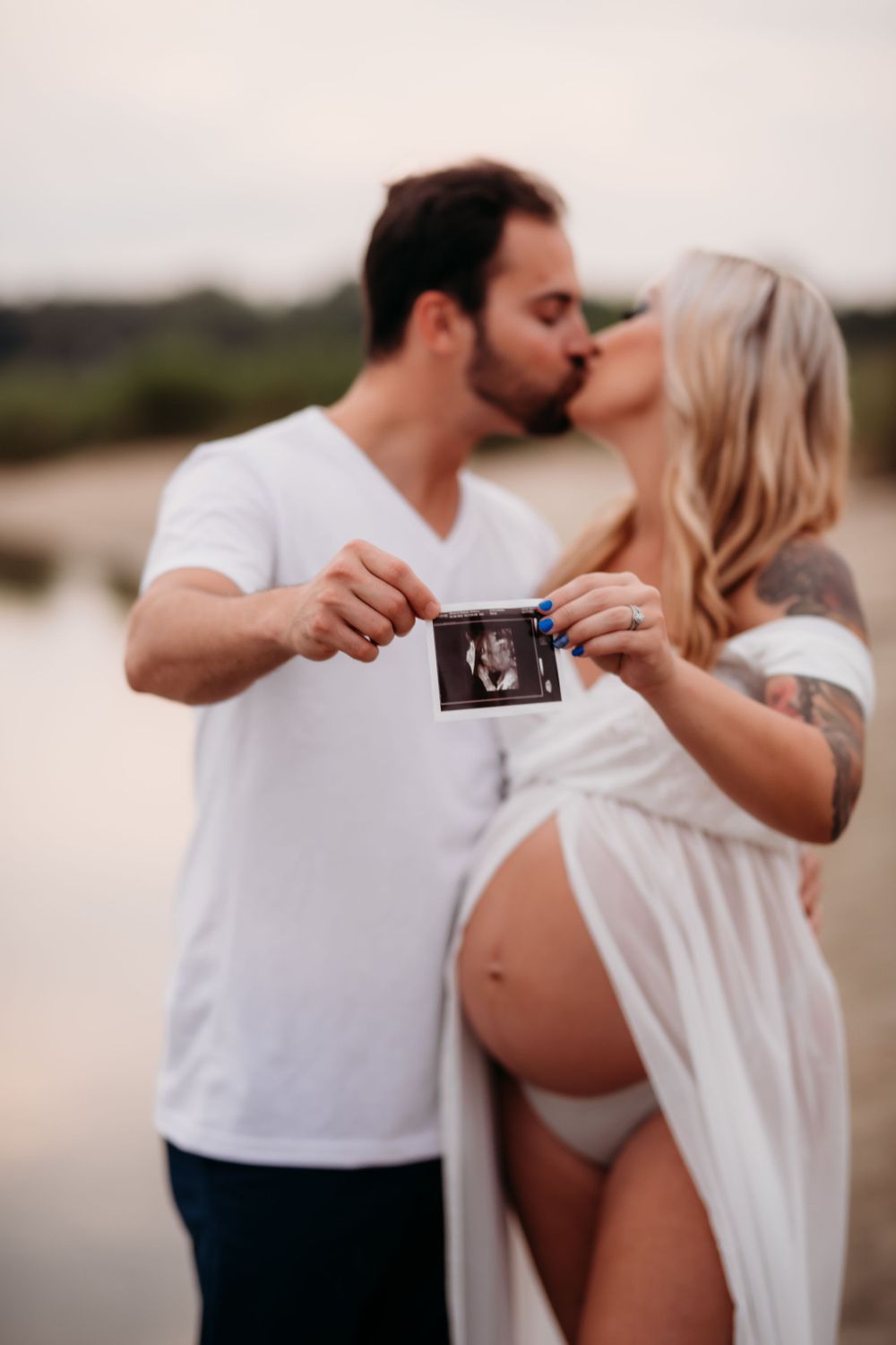 Couple kisses while holding ultrasound photo on beach during maternity photoshoot.