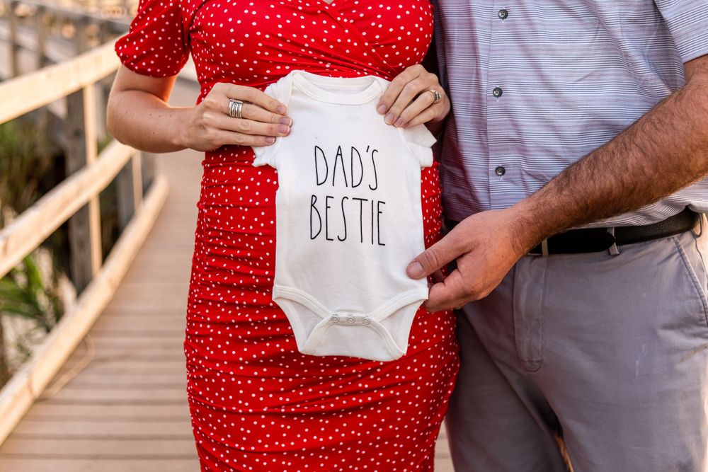 A couple holds up a white onesie with 'Dad's Bestie' text while standing on a wooden boardwalk.