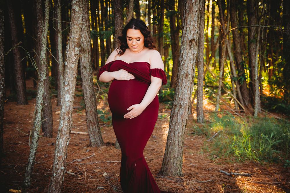 Maternity photo in a burgundy gown among tall forest trees at sunset.