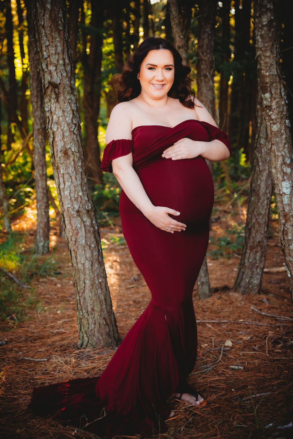 Beautiful maternity session in a burgundy off-shoulder gown surrounded by forest trees.