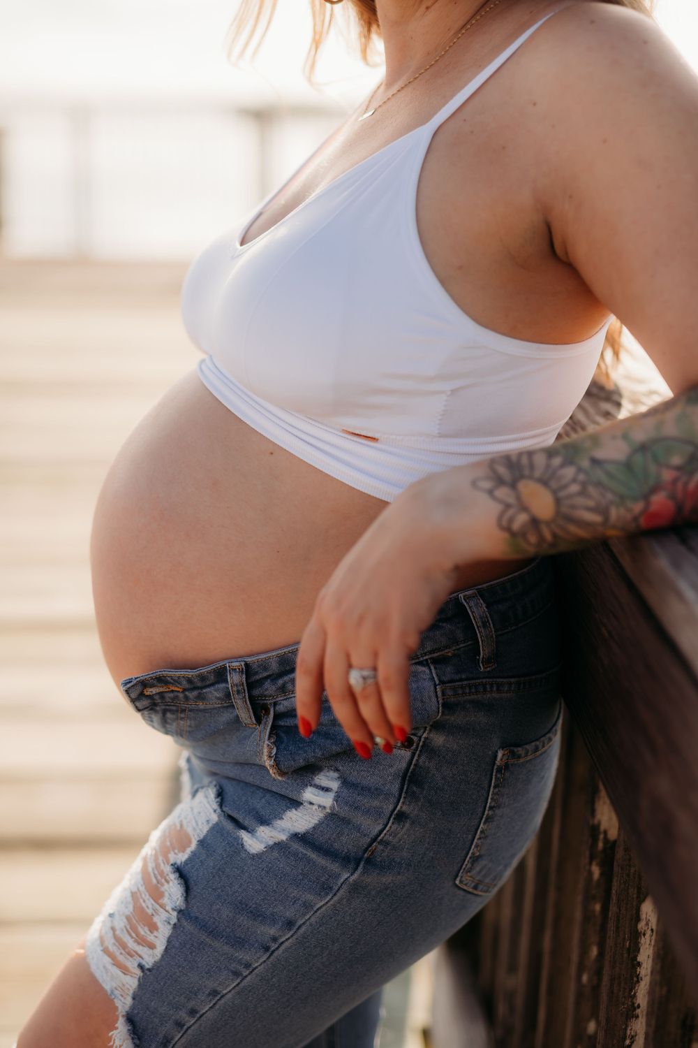 Side view of pregnant woman in white top and ripped jeans standing against sunset light.