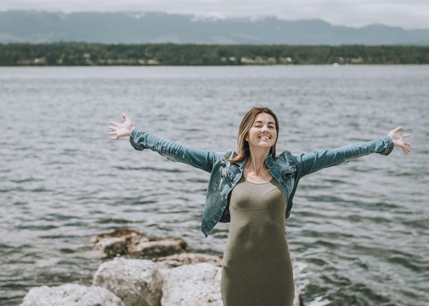 Person with outstretched arms stands joyfully on rocky shoreline against mountain lake backdrop.