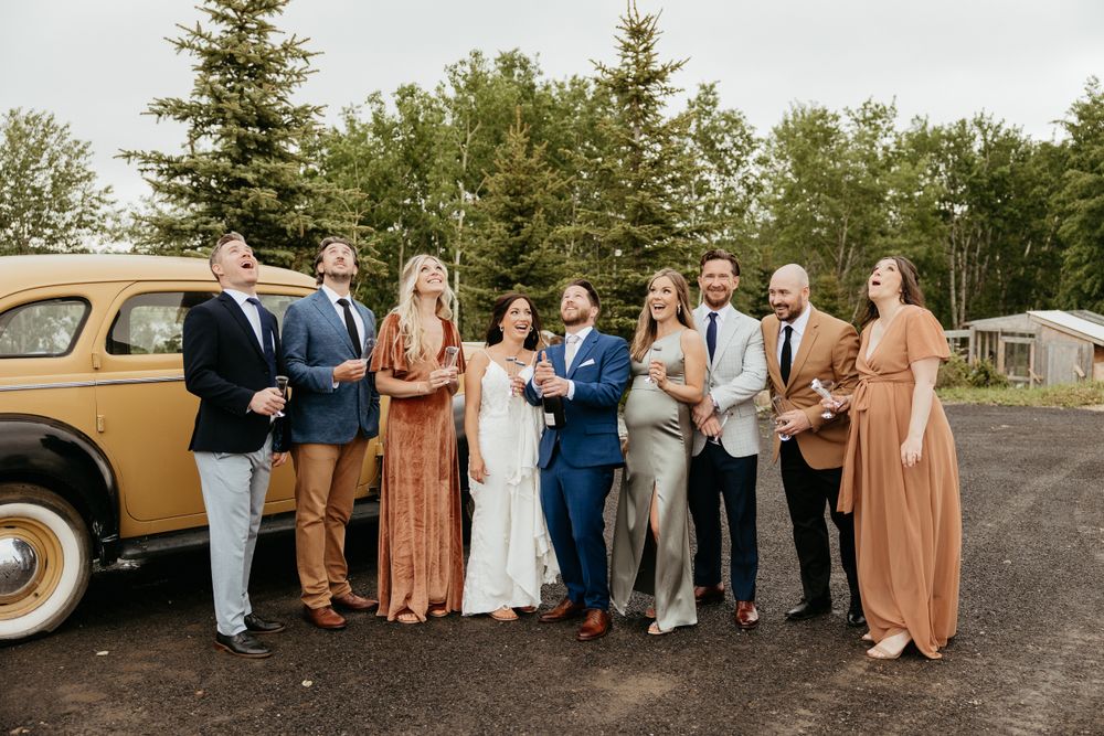 Wedding party members pose together in front of a vintage yellow vehicle outdoors.