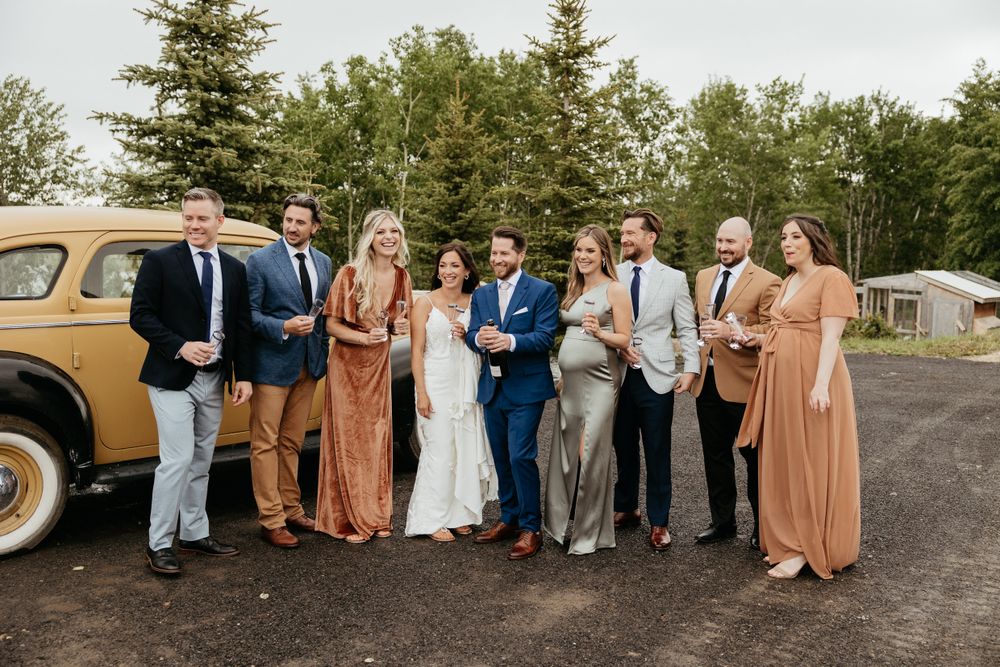 Wedding group photo with bridal party wearing earth tones standing by a classic car.