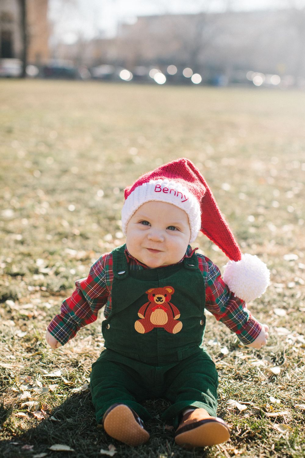 Baby Benny in a Santa hat and green teddy bear overalls sitting on grass outdoors for a Christmas photo.