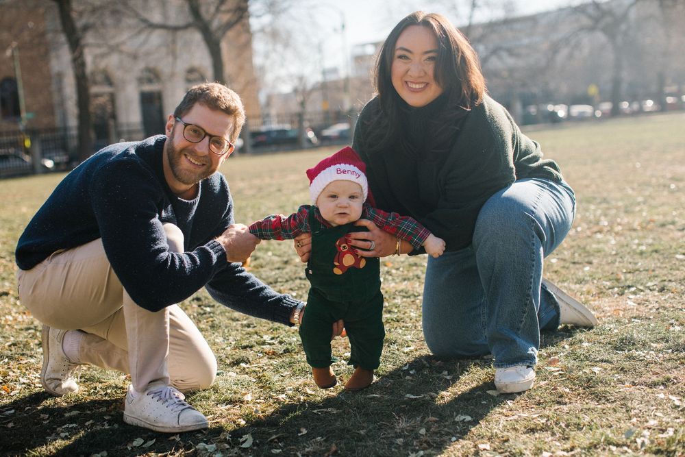 A young family poses outdoors on grass, parents helping their baby in a Christmas outfit and Santa hat stand up.